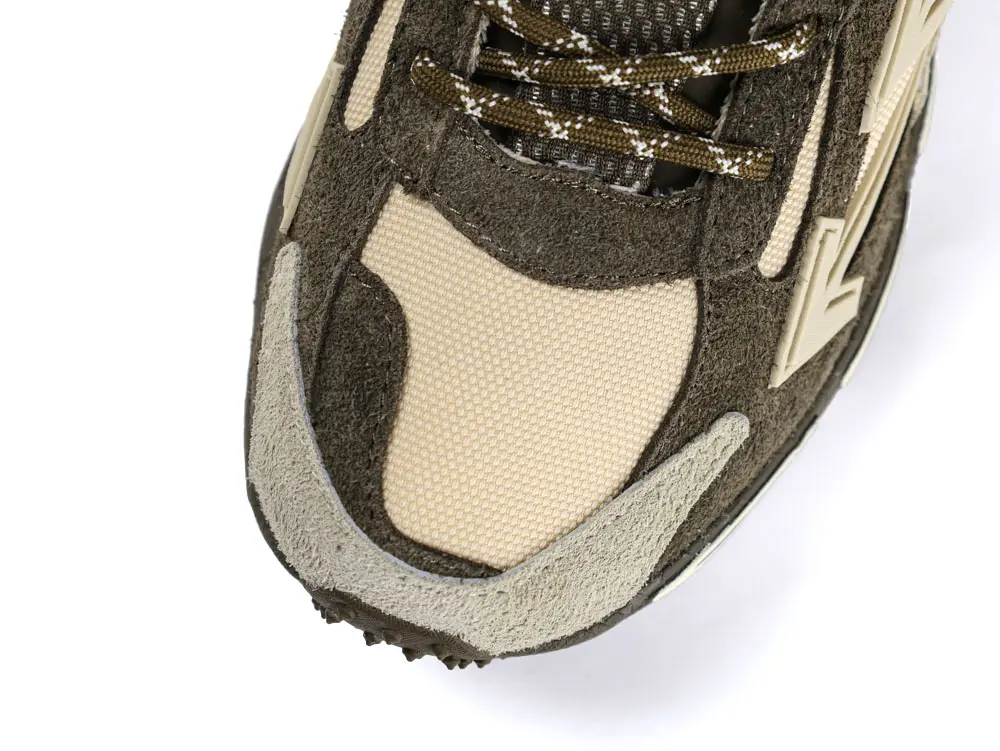 Close-up of a sneaker focusing on the toe area. The shoe features a combination of suede and mesh materials in dark green and beige colors. The laces are dark with light speckles, and part of the shoe's logo is visible on the side. The texture and stitching details of the shoe are prominent.