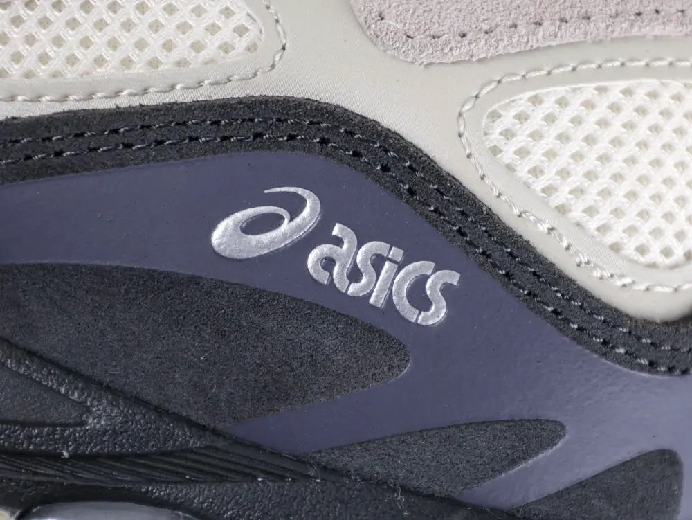 Close-up of a sneaker, featuring gray leather, mesh panels, and an "Asics" logo.